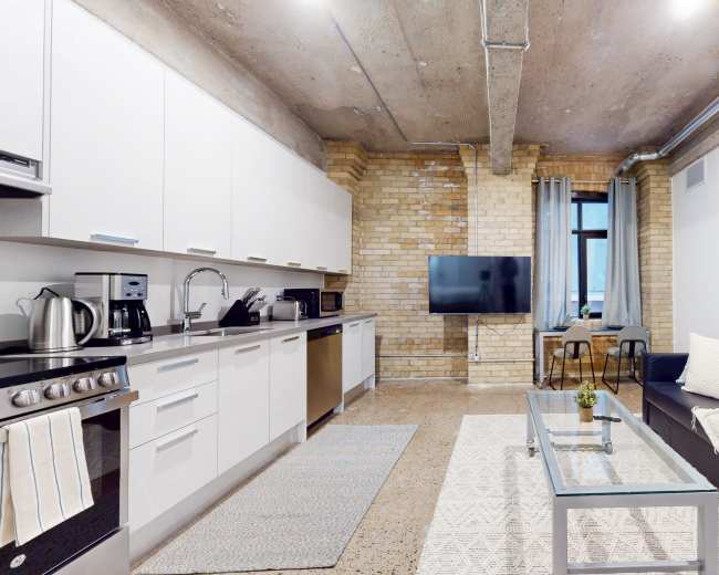 The image shows a modern apartment interior featuring a kitchen area on the left, a living space with a black sofa and coffee table in the center, and a television mounted on a brick wall.