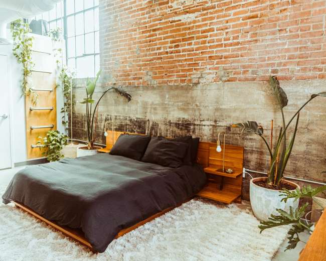 The image depicts a minimalistic bedroom featuring a low wooden bed with a black duvet, surrounded by potted plants and a textured brick wall.