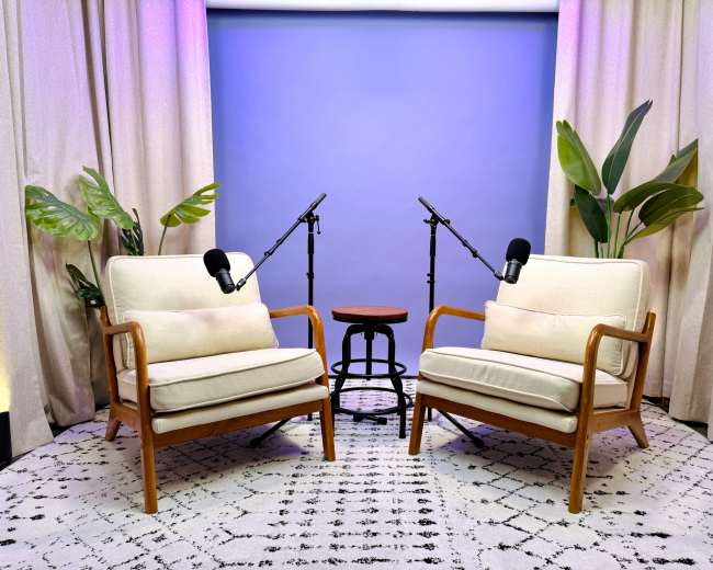 The image shows two cushioned chairs on a patterned rug, positioned opposite each other with a small table and microphones set up between them, surrounded by plants and draped curtains.