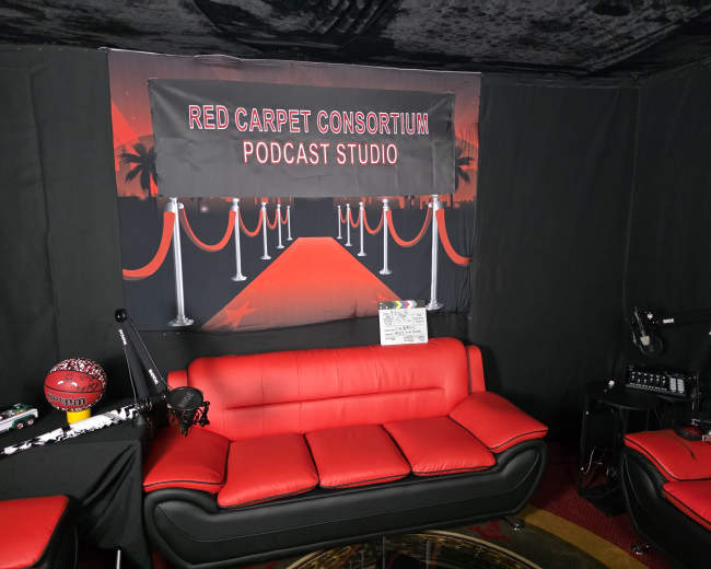 The image shows a podcast studio with red couches, a backdrop featuring a red carpet theme, and recording equipment.