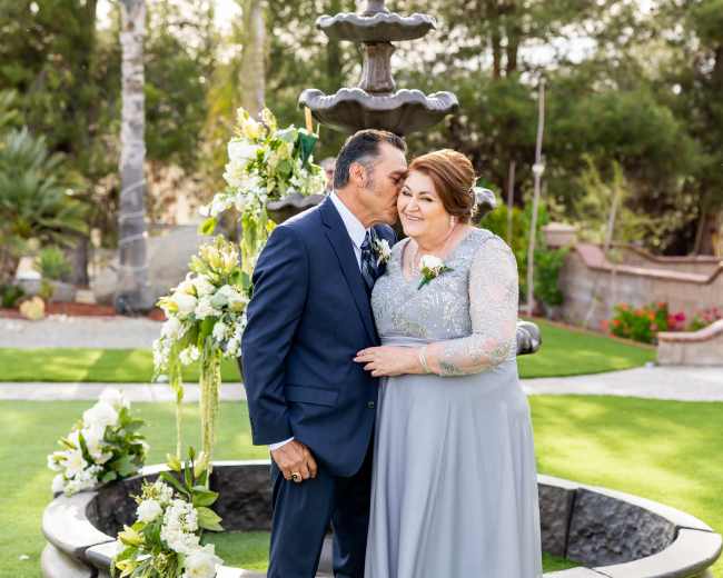 A couple stands beside a decorative fountain, surrounded by greenery and floral arrangements.
