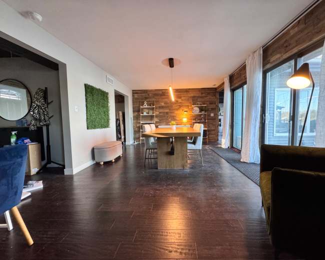 The image shows a modern living space featuring a dining table in the center, surrounded by chairs, with large windows and a wood-paneled accent wall.