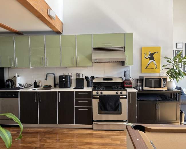 The image shows a modern kitchen with green upper cabinets, dark lower cabinets, and various appliances including a stove, microwave, and coffee maker.