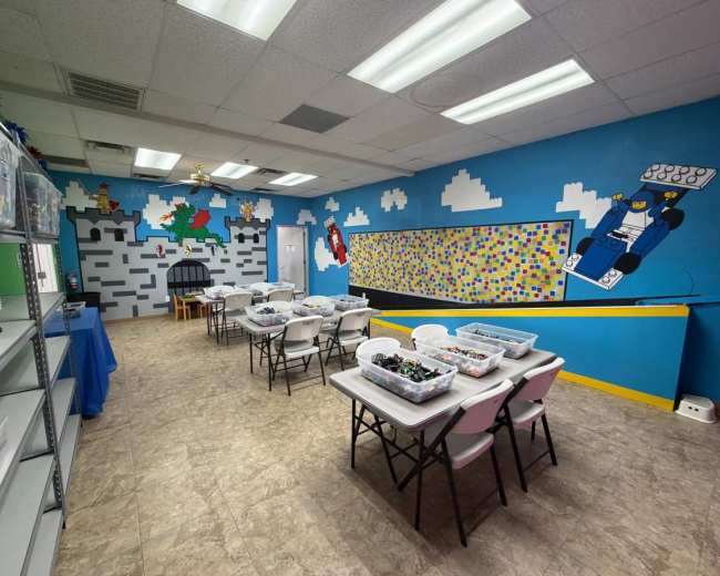 The image shows a bright classroom with several tables arranged for activities, surrounded by colorful murals of a castle and clouds on the walls.