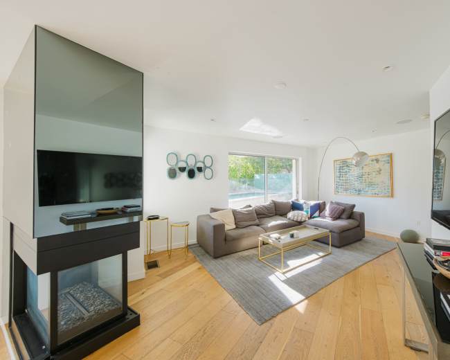 The image shows a modern living room with a gray sectional sofa, a coffee table on a rug, and large windows allowing natural light to enter.