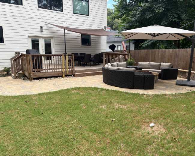 The image shows a backyard with a paved seating area featuring a circular lounge set and a large umbrella, next to a wooden deck furnished with chairs and a shaded canopy.