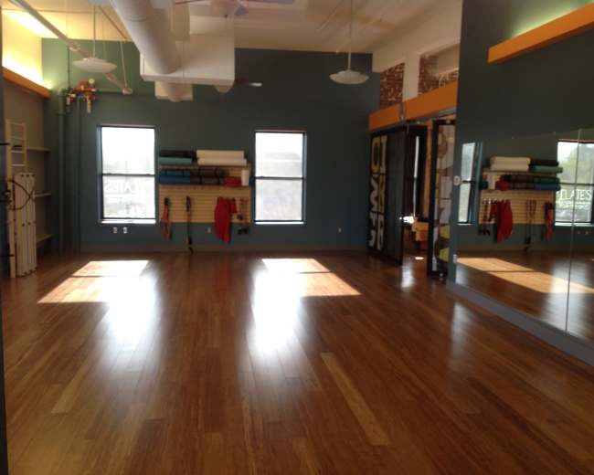 The image shows a spacious wooden-floored studio with large windows, mirrors, and exercise equipment along the walls.