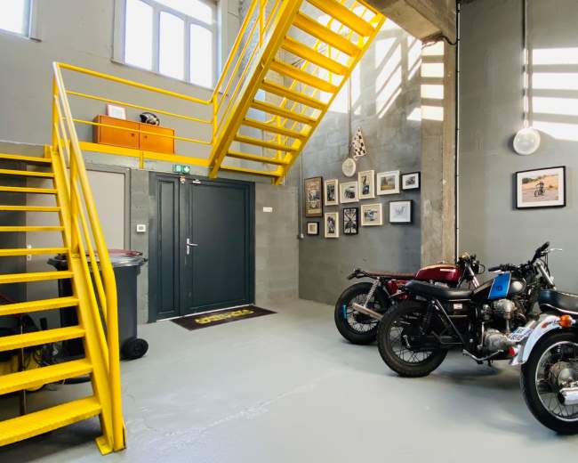 The image shows a modern garage space featuring three motorcycles parked near a gray wall, with a bright yellow staircase leading to an upper level.