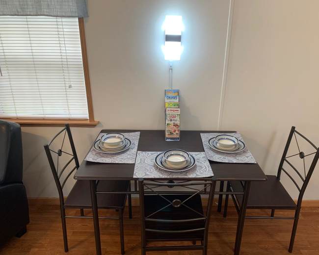 A small dining table set with four chairs, placemats, and plates, is positioned against a wall with a lamp and a display of snack boxes nearby.