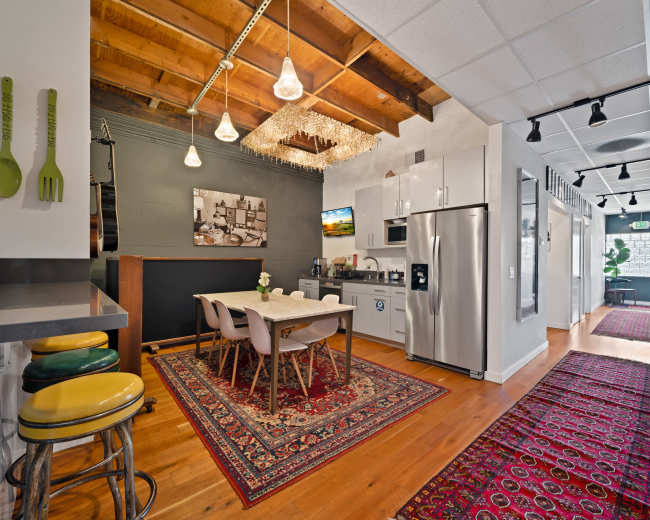 The image shows a well-lit kitchen and dining area with a wooden table, modern appliances, and a hallway leading to additional rooms, featuring a decorative rug.