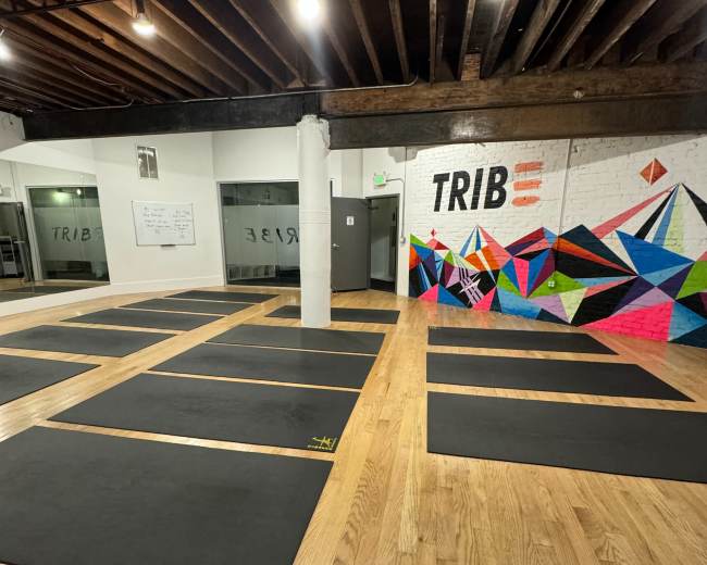 The image shows a spacious yoga studio with several black mats arranged neatly on a wooden floor, and a colorful geometric mural on the wall.