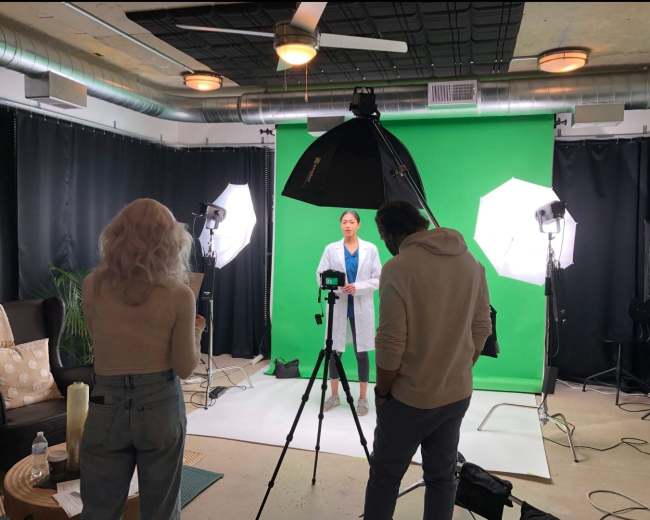 A woman dressed in a lab coat stands in front of a green screen while two individuals operate a camera and lighting equipment in a studio setting.