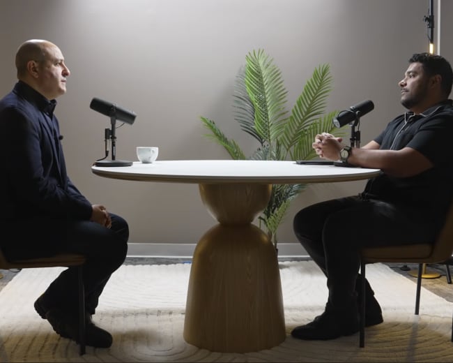 Two men are seated at a round table in a well-lit room, each positioned in front of a microphone, with a plant in the background.