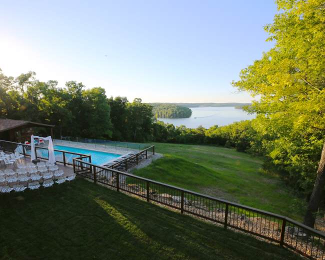 A swimming pool with lounge chairs overlooks a scenic lake surrounded by trees.