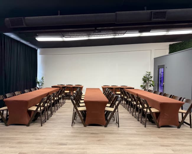 The image shows a spacious meeting room with two rows of long tables covered in orange tablecloths, arranged in a U-shape, with black chairs around them.