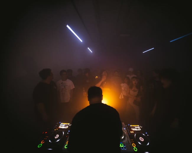 A DJ stands at a control table in a dimly lit room, with a crowd of people dancing in the background illuminated by soft, colorful lights.