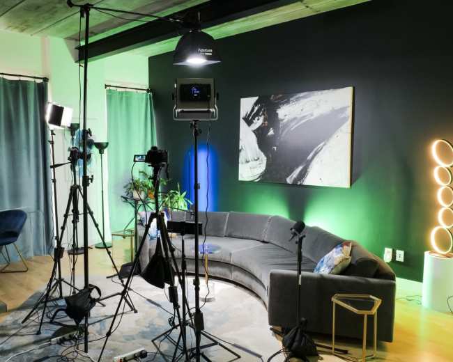The image shows a recording or photography setup featuring a circular sofa, professional lighting equipment, cameras on tripods, and a control station with monitors in a studio-like environment.