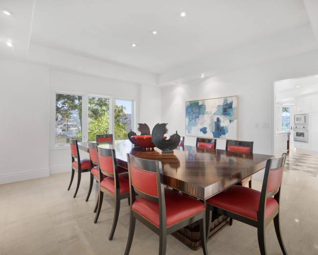A modern dining room features a large wooden table surrounded by red-upholstered chairs, with artwork on the walls and windows letting in natural light.