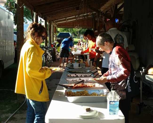 A group of people gather around a long table under a covered area, serving and enjoying various dishes during a community event.