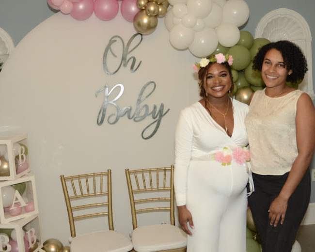 Two women stand in front of a decorated wall with the words "Oh Baby," surrounded by balloons and a display of blocks.