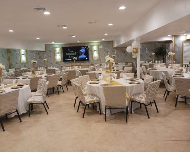 The image shows a banquet hall with round tables dressed in white tablecloths and gold accents, surrounded by upholstered chairs, and featuring wall decorations and a large screen at the front.