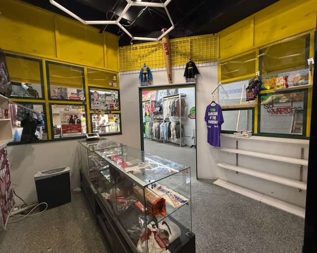The image shows the interior of a retail store featuring a glass display case, shelves stocked with various items, and clothing hung on racks along the walls.