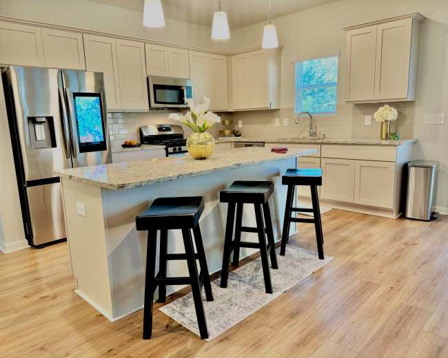 A modern kitchen features a central island with three black stools, stainless steel appliances, and light-colored cabinetry.