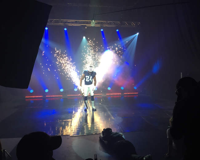 A football player in a blue jersey with the number 24 stands on a reflective stage illuminated by bright lights and sparks.