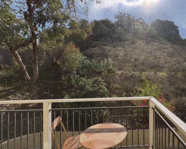 A small balcony with a round wooden table overlooks a hillside covered in sparse vegetation and trees under a partially cloudy sky.