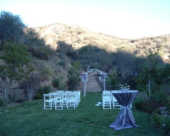 A decorated outdoor wedding ceremony setup features rows of white chairs and a floral arch on a grassy area overlooking a hillside.