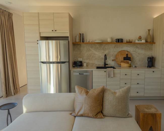 A modern kitchen features wooden cabinetry, a stainless steel refrigerator, and a stone backsplash, with a cozy seating area visible in the foreground.