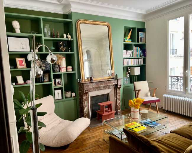 The image shows a stylish living room with green shelving, a large mirror above a marble fireplace, and a cozy seating area that includes a modern chair and a lounge chair.