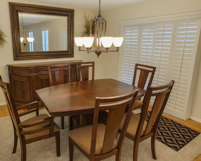 A wooden dining table surrounded by six chairs is positioned in a well-lit room with a mirror and a sideboard against the wall.