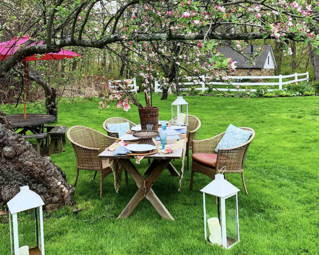 A table set for a meal sits under a blossoming tree in a green garden, with wicker chairs and lanterns nearby.