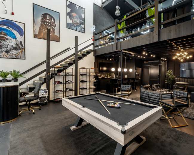 The image shows a modern interior space featuring a billiard table at the center, with a staircase, a lounge area, and various artworks on the walls.