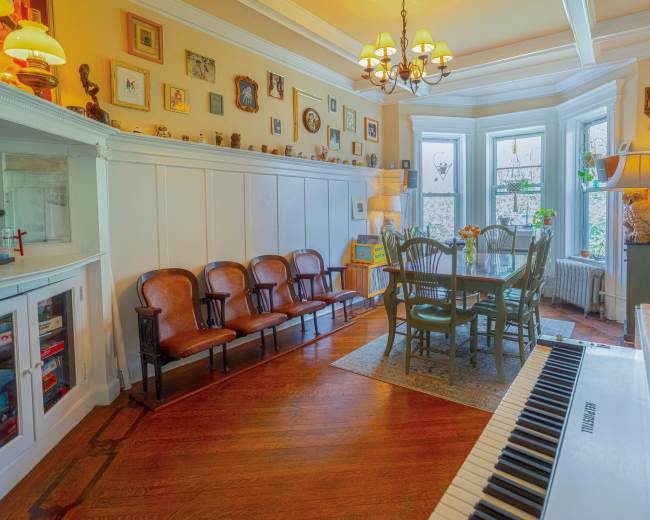 A cozy room features a dining table surrounded by chairs, with a row of vintage armchairs and walls adorned with framed pictures.