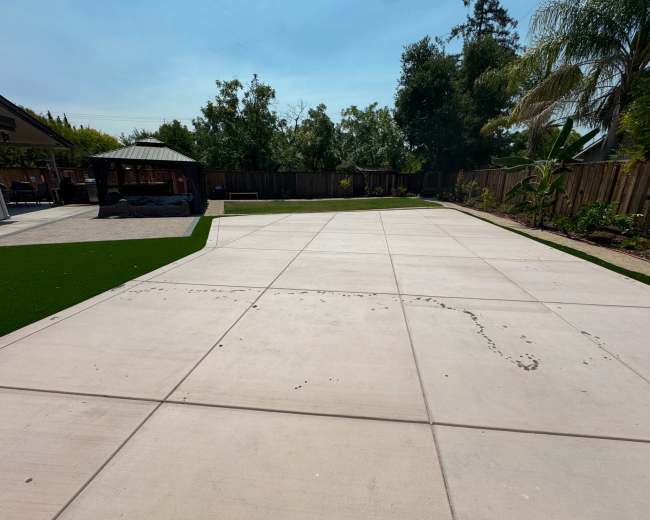 The image shows a large, empty concrete patio with a patch of artificial grass on one side, surrounded by trees and a fence.