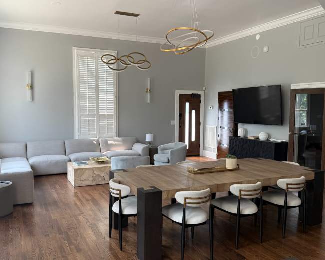 The image shows a modern living room and dining area featuring a gray sectional sofa, a wooden dining table with chairs, and contemporary light fixtures.