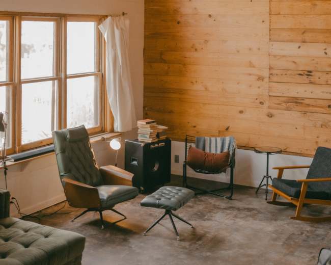 The image shows a spacious interior of a room with wooden walls, large windows, and several chairs arranged in a minimalist style.