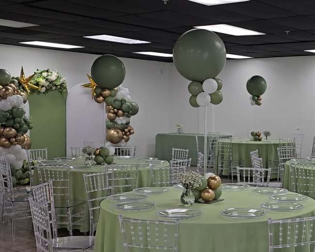 The image shows a banquet hall decorated with green and gold balloons, circular tables covered in green tablecloths, and transparent Chiavari chairs.