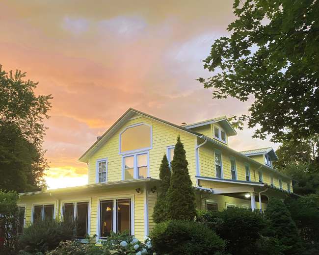 A yellow house with multiple windows is framed by greenery, under a sky illuminated by a colorful sunset.