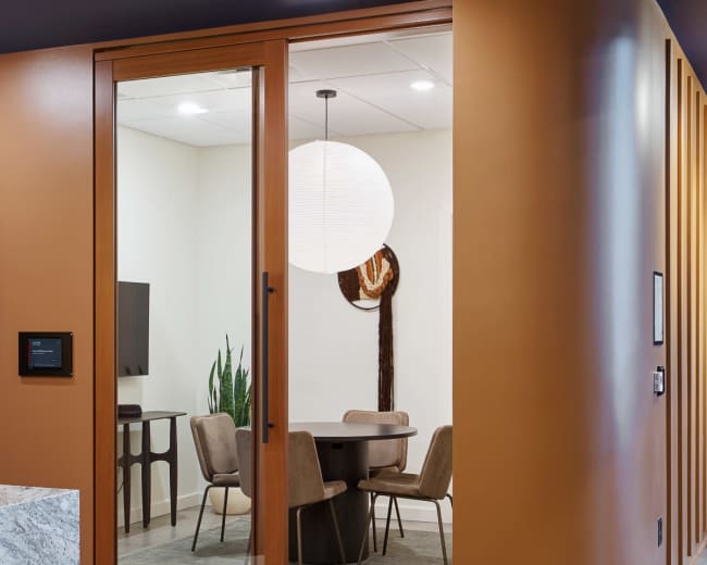 A modern conference room with a circular table and upholstered chairs is visible through a glass door, set against warm, orange-toned walls.