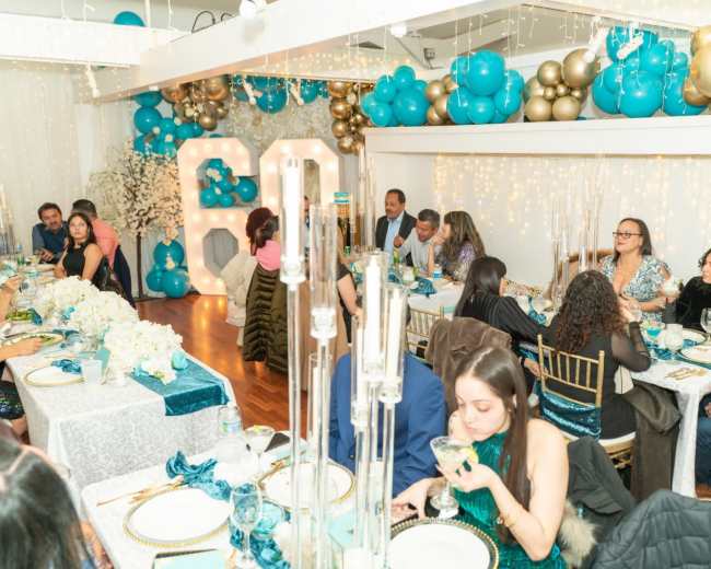 A decorated banquet room hosts a gathering with guests seated at elegantly set tables, surrounded by balloons and floral arrangements.