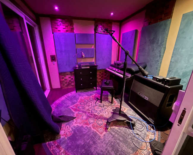 The image shows a sound recording studio with acoustic panels on the walls, a microphone on a stand, and various musical equipment.