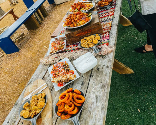 A wooden table is filled with various platters of food, including fried items, salads, and sauces, set in an outdoor area with benches nearby.