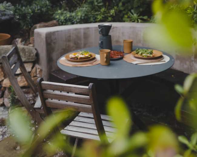 A table set with two plates of food and coffee cups is situated in a garden surrounded by greenery.