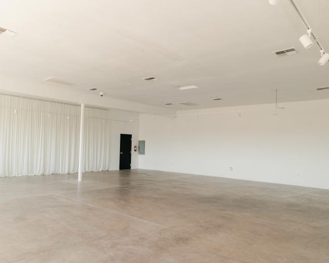 The image shows a spacious, empty room with concrete flooring, white walls, and a large curtain-covered window.