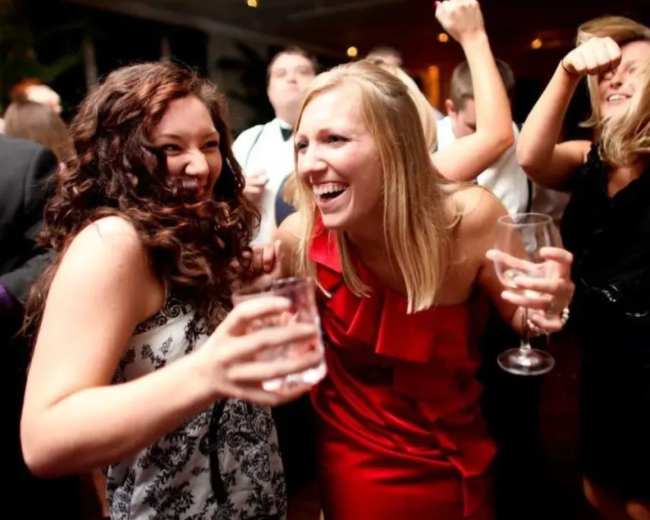 Two women are smiling and holding drinks while dancing at a lively party, surrounded by other guests.