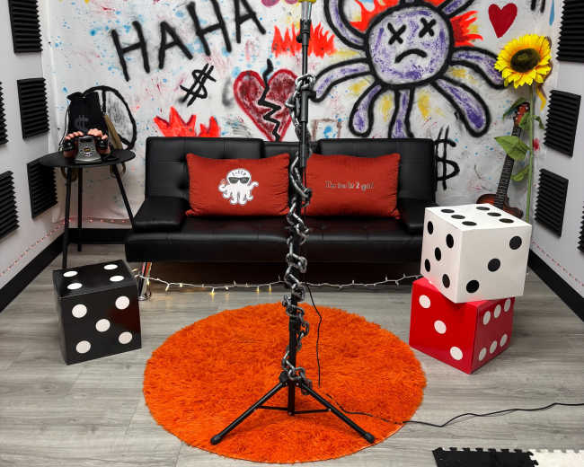 The image shows a colorful room with a black sofa, a bright orange rug, decorative dice, and a background featuring graffiti art with symbols and messages.