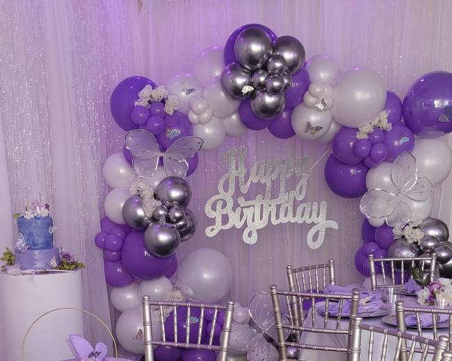 A birthday party setup with a table adorned in purple and silver decorations, featuring a cake and a backdrop of balloons.